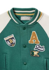 Veste varsity verte avec des manches crème, ornée de patchs brodés en jaune, marine et blanc. Comprend des boutons-pression et un col rayé.