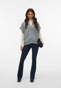 Grå stickad väst med korta ärmar, dragkedja fram och kort nederkant, kombinerad med en ljus polotröja och mörka utsvängda jeans, håller en svart kuvertväska.