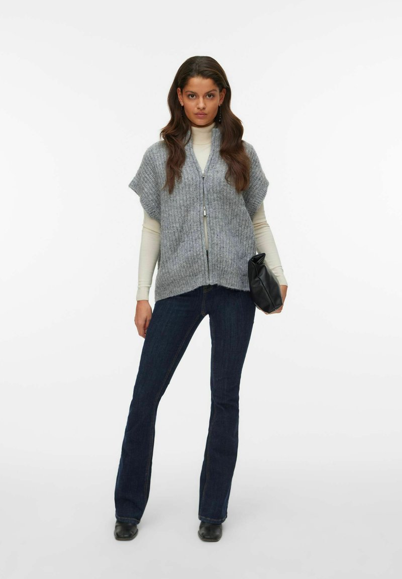 Grå stickad väst med korta ärmar, dragkedja fram och kort nederkant, kombinerad med en ljus polotröja och mörka utsvängda jeans, håller en svart kuvertväska.