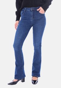 Jeans flare in denim blu scuro, con vita a media altezza, cinque tasche e una texture liscia. Abbinati a tacchi in pelle verniciata nera.