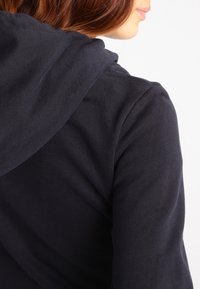 Kvinna med rött hår iförd svart hetjacka, synlig från baksidan och sidan, mot en enfärgad ljus bakgrund.