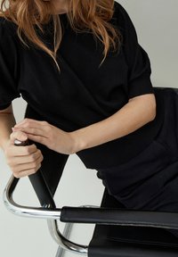 Femme aux cheveux ondulés assise de profil sur une chaise en cuir noir et chrome, portant un haut noir à manches courtes et un pantalon, les mains reposant sur l'accoudoir de la chaise.