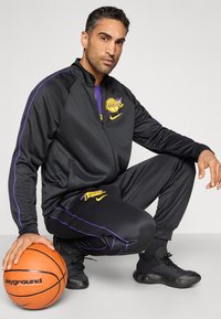 Μαύρο αθλητικό φόρμα με μωβ λεπτομέρειες και λογότυπο Lakers. Το μοντέλο κρατάει πορτοκαλί μπάλα μπάσκετ και φοράει μαύρα αθλητικά παπούτσια.