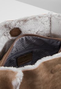 Taske i ruskind i cognacfarve med blødt gråt pelsfor. Har en lynlåslomme og et mærket lædermærke. Enkelt design med rund knaplukning.