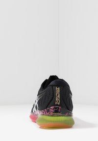 Zapatilla deportiva negra con parte superior texturizada, acentos plateados y una suela multicolor que presenta detalles en rosa, verde y naranja.