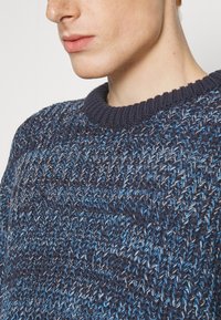 Námořnický modrý pletený svetr s texturovaným vzorem, který zahrnuje různé odstíny modré a bílé. Silný žebrovaný výstřih dodává kontrast.