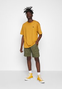 Camiseta oversized color mostaza con logo negro, pantalones cortos cargo verdes y zapatillas altas amarillas, de pie contra un fondo blanco.