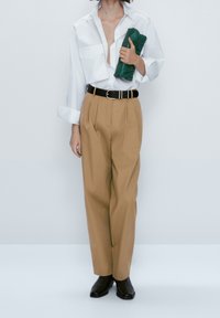 Vit långärmad skjorta, beige veckade byxor, svart bälte och svarta ankelstövlar. Håller en grön clutchväska. Minimalistisk design.