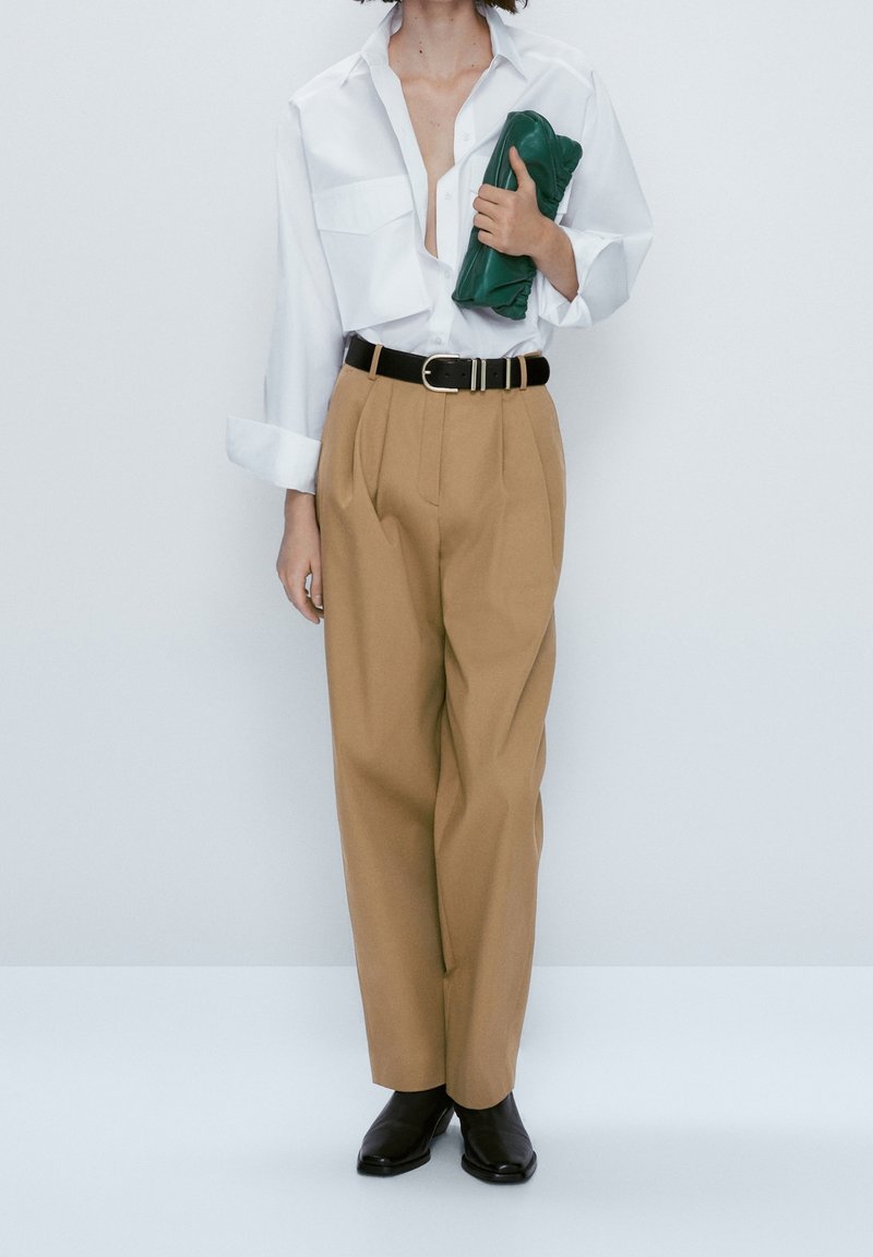 Vit långärmad skjorta, beige veckade byxor, svart bälte och svarta ankelstövlar. Håller en grön clutchväska. Minimalistisk design.