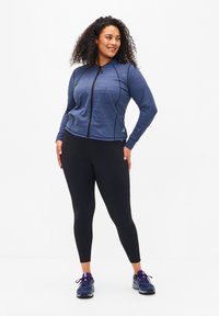 Dunkelblaues sportliches Jacket mit Reißverschluss, strukturierter Stoff und taillierter Form, kombiniert mit schwarzen Leggings und dunklen Turnschuhen.