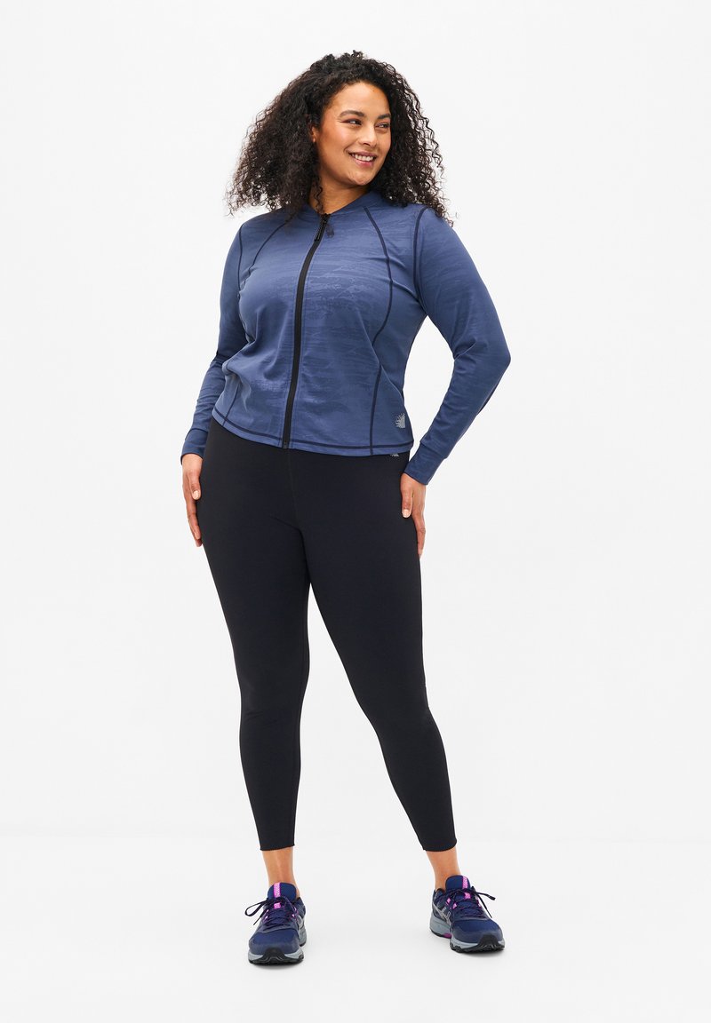 Dunkelblaues sportliches Jacket mit Reißverschluss, strukturierter Stoff und taillierter Form, kombiniert mit schwarzen Leggings und dunklen Turnschuhen.