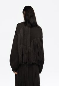 Jersey negro de gran tamaño con largas borlas en la parte trasera, tejido texturizado y mangas abullonadas. Transición suave de las áreas sólidas a las áreas con flecos.