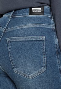 Dark blue denim jeans with a mid-rise fit, featuring a black DRDENIM label on the waistband and standard back pockets with stitching detail.