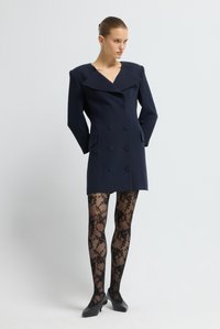 Cappotto navy doppiopetto con ampio colletto, tasche laterali e spalle strutturate, abbinato a collant neri a fantasia e tacchi neri.