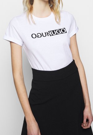 Nainen, jolla on yllään valkoinen T-paita, jossa on käännetty musta "HUGO"-logo, sekä korkeavyötäröinen musta hame, tasaisella vaalealla taustalla.