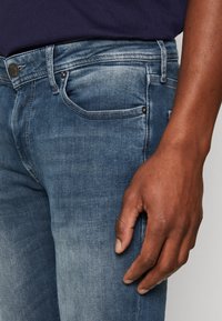 Jeans de mezclilla azul oscuro con un diseño clásico de cinco bolsillos, textura ligeramente deslavada y detalles de costura en contraste.