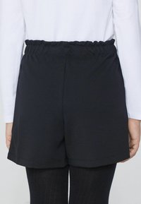 Czarne szorty na elastycznej talii o gładkiej fakturze i przeszywanych brzegach, w połączeniu z czarnymi legginsami. Widok z tyłu nie pokazuje dodatkowych szczegółów.