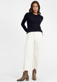 Schwarzer strukturierter Pullover, weiße weit geschnittene Hose mit fransigen Säumen und braune Stiefeletten mit Schlangenprint. Sauberes, modernes Outfit-Design.