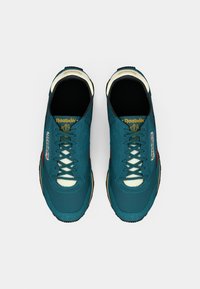 Tênis atléticos em azul-petróleo com detalhes em creme e solas de borracha pretas. Possui atacadores e logótipos na língua e nas laterais. Textura suave.