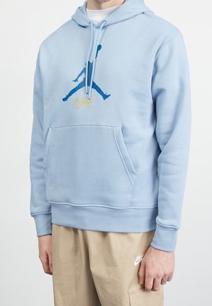 Person wearing light blue hoodie with a blue silhouette logo and yellow "Flight" text, paired with beige Nike pants.