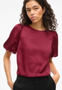 Haut en satin bordeaux avec manches courtes bouffantes et encolure ronde. Texture lisse et silhouette cintrée associées à un pantalon foncé.