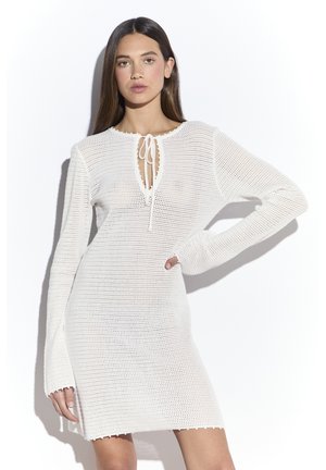Donna con lunghi capelli castani che indossa un vestito bianco a maniche lunghe in maglia con un laccio al collo, che posa con una mano sul fianco.