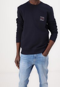 Persona che indossa una felpa blu navy con un piccolo logo Tommy Hilfiger sul petto e jeans azzurri chiari, con una mano in tasca.