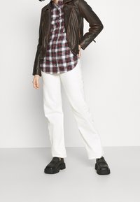 Bruine leren motorjack, rood en zwart geruite blouse, witte wijde broek en zwarte platform schoenen. Eenvoudige texturen en gelaagd uiterlijk.