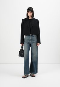 Kvinna som bär en kort svart kavaj, svart topp, vida blå jeans, ormskinnsandaler och håller en liten svart handväska.