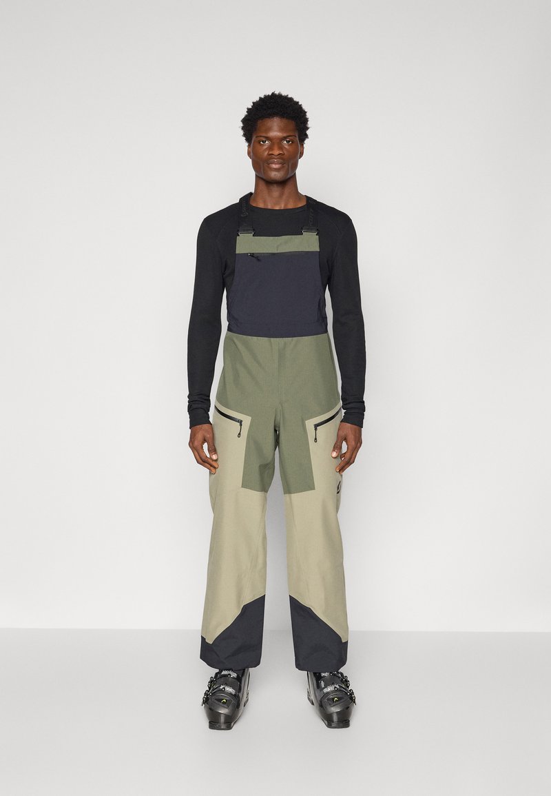 Kombinezon narciarski w zielonym i beżowym kolorze, posiada odpinane ramiączka, kieszenie na zamek oraz czarne akcenty. Materiał wydaje się trwały z teksturowanym wykończeniem.