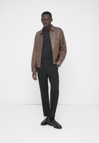 Veste en cuir marron avec un t-shirt noir à col rond, assortie à un pantalon noir et des chaussures noires. Texture lisse et détails avec fermeture éclair.