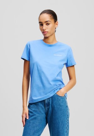 Blå bomulds t-shirt med rund hals og korte ærmer, prydet med hvidt logo tekst på venstre bryst. Sammen med blå denim jeans.