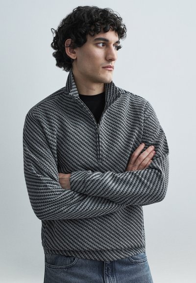 Pull-over à fermeture éclair gris avec un motif en chevron, manches longues et col montant, porté sur une chemise noire et associé à un jean bleu.