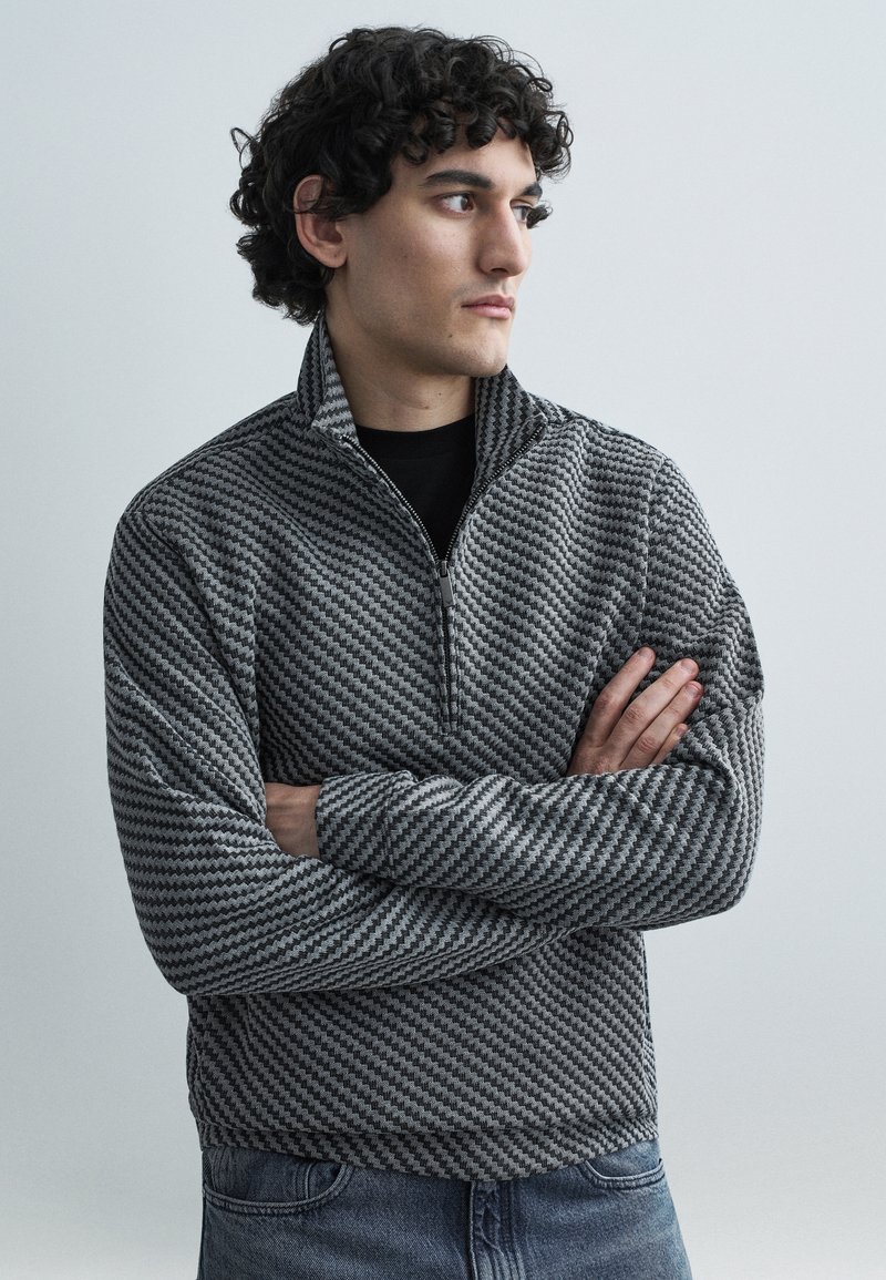 Šedá pulóverová bunda na zips so vzorom šípok, dlhými rukávmi a vysokým golierom, nosená cez čierne tričko a kombinovaná s modrými džínsami.