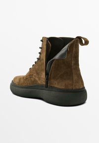 Bottes chelsea en suède marron avec une semelle en caoutchouc noir, présentant une fermeture éclair sur le côté, un laçage à l'avant et une languette au niveau du talon.