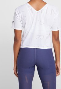 Vit kortärmad träningsskjorta med V-ringning och ett mönster av perforerad text över hela ytan. I kombination med mörk lila leggings med ett liknande mönster.