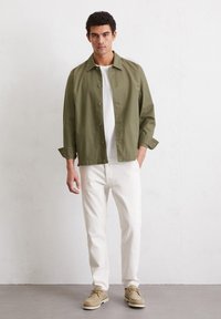 Olijfgroene lichtgewicht jas, met een knoopsluiting aan de voorkant en een kraag, gecombineerd met een wit t-shirt en witte broek. Schoenen zijn beige suède met veters.