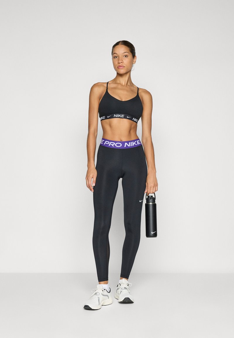 Soutien-gorge de sport noir avec le logo Nike, leggings taille haute noirs avec une bandeau Nike violette, tenant une bouteille d'eau noire, baskets blanches.