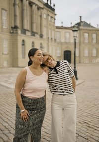 Two women stand close together, sharing a smile. One wears a pink sleeveless top and patterned skirt; the other a striped polo and white pants.