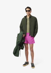 Giacca leggera verde, polo oliva, pantaloni corti rosa e scarpe da ginnastica grigie. Indossa occhiali da sole, tiene la giacca, con maniche corte visibili.