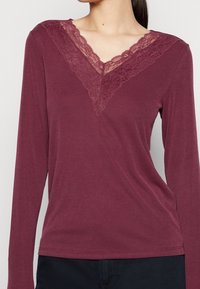 Burgunderfarbener Langarmtop mit gerippter Textur und Spitzenbesatz am V-Ausschnitt. Das Shirt hat eine lockere Passform und eine glatte Oberfläche.