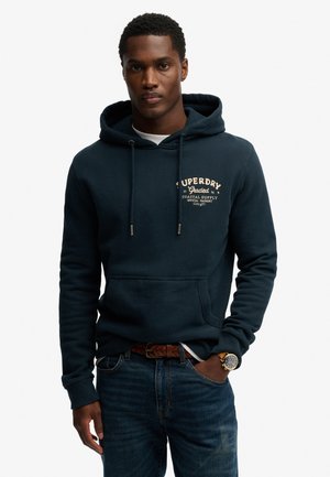 Homme portant un sweat à capuche Superdry bleu marine avec poche avant, logo imprimé, ceinture tressée marron, jean foncé et montre-bracelet dorée, debout avec la main dans la poche.