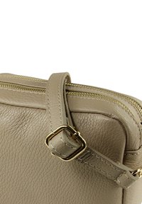 Beige leren handtas met een getextureerd oppervlak, gouden ritssluiting en verstelbare riem met een rechthoekige gesp. Compact ontwerp, minimalistische details.