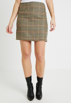 Femme portant une mini-jupe marron et noire à motif pied-de-poule, associée à des bottines en cuir noir, debout devant un arrière-plan clair uni.