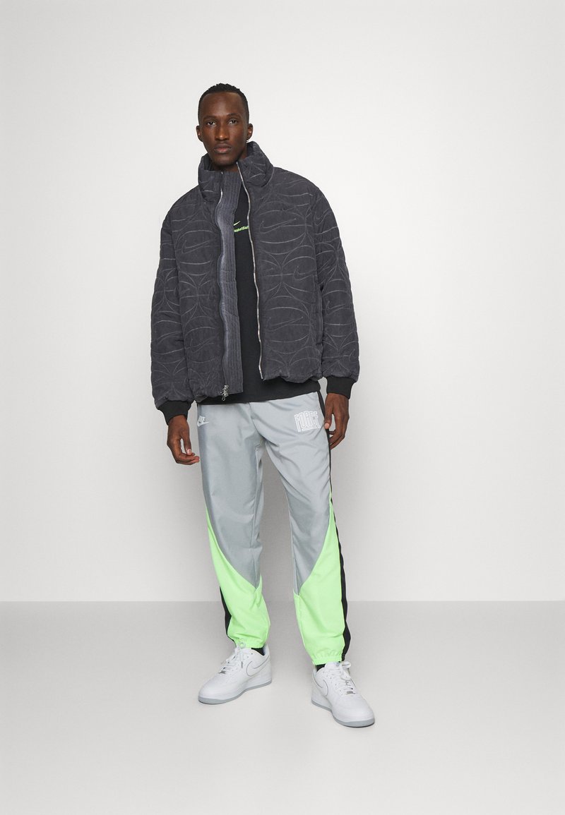 Uomo in piedi che indossa una giacca trapuntata scura, una maglietta nera, pantaloni da jogging grigi e verde neon, e sneakers bianche su uno sfondo neutro.