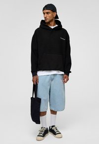 Sweat-shirt à capuche oversized noir avec une poche avant, shorts en denim bleu clair, chaussettes blanches et baskets noires tenant un grand sac en toile noir.