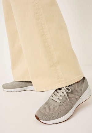 Ben i beige bukser med vide ben og lysegrå strikkede sneakers med hvide såler på en lys baggrund.