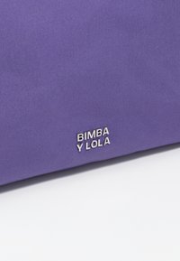 Violetti olkalaukku, jossa on sileä pinta ja hopeiset logoyksityiskohdat. Ominaisuuksiin kuuluu jämäkkä muotoilu ja tilava avaus helppoa käyttöä varten.