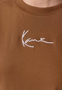 Brown short-sleeve t-shirt with a crew neckline featuring white embroidered text that reads "Kani." Material appears soft and textured.