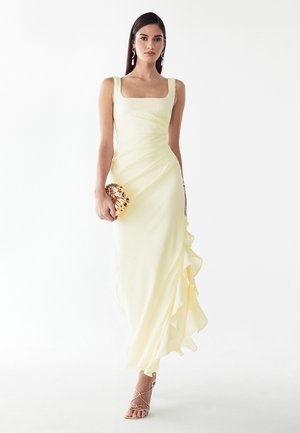 Mujer con vestido sin mangas de color amarillo pálido con detalle de volantes, sosteniendo un bolso de mano dorado en forma de concha y luciendo tacones dorados con tiras.